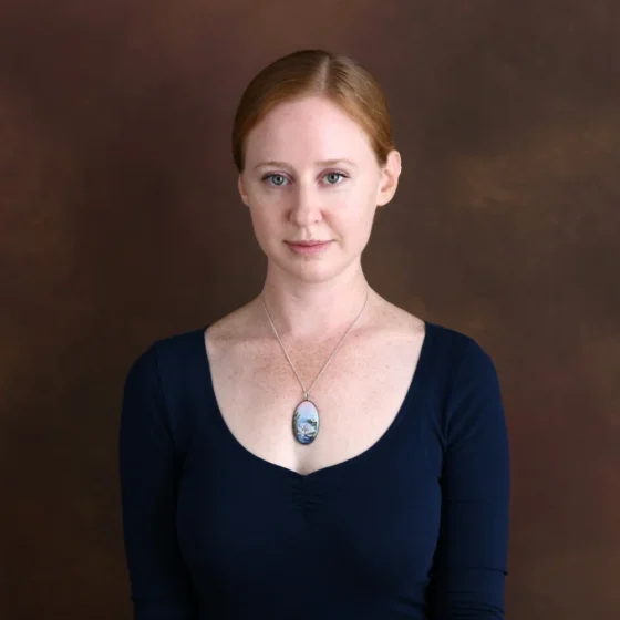 A woman with red hair pulled back and blue eyes. She wears a black shirt and a large, blue pendant with flowers on it.