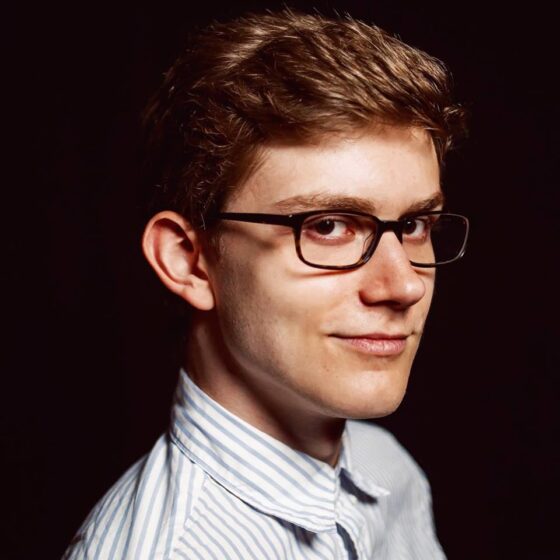 A man with straight, short, dirty-blonde hair and brown eyes, smiling. He wears a blue-and-white-striped shirt and black, rectangular glasses.