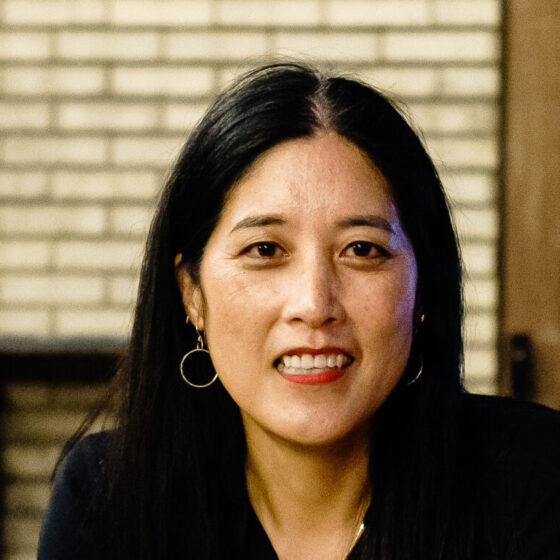 An Asian woman with a dark V-neck shirt and earrings. She's in front of a white brick wall.