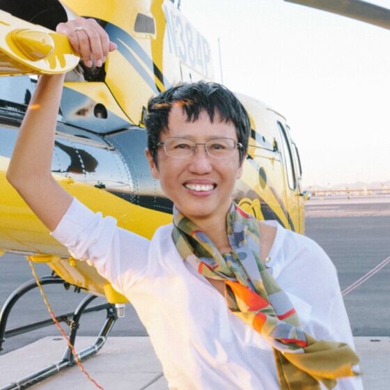 A woman with a straight, black, pixie cut and dark eyes, smiling. She leans against the wing of a yellow plane, wearing a white shirt and a multi-colored scarf.