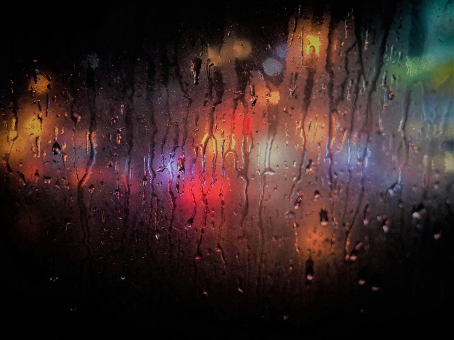 Different colored lights shine in the night behind a foggy, rain-covered window