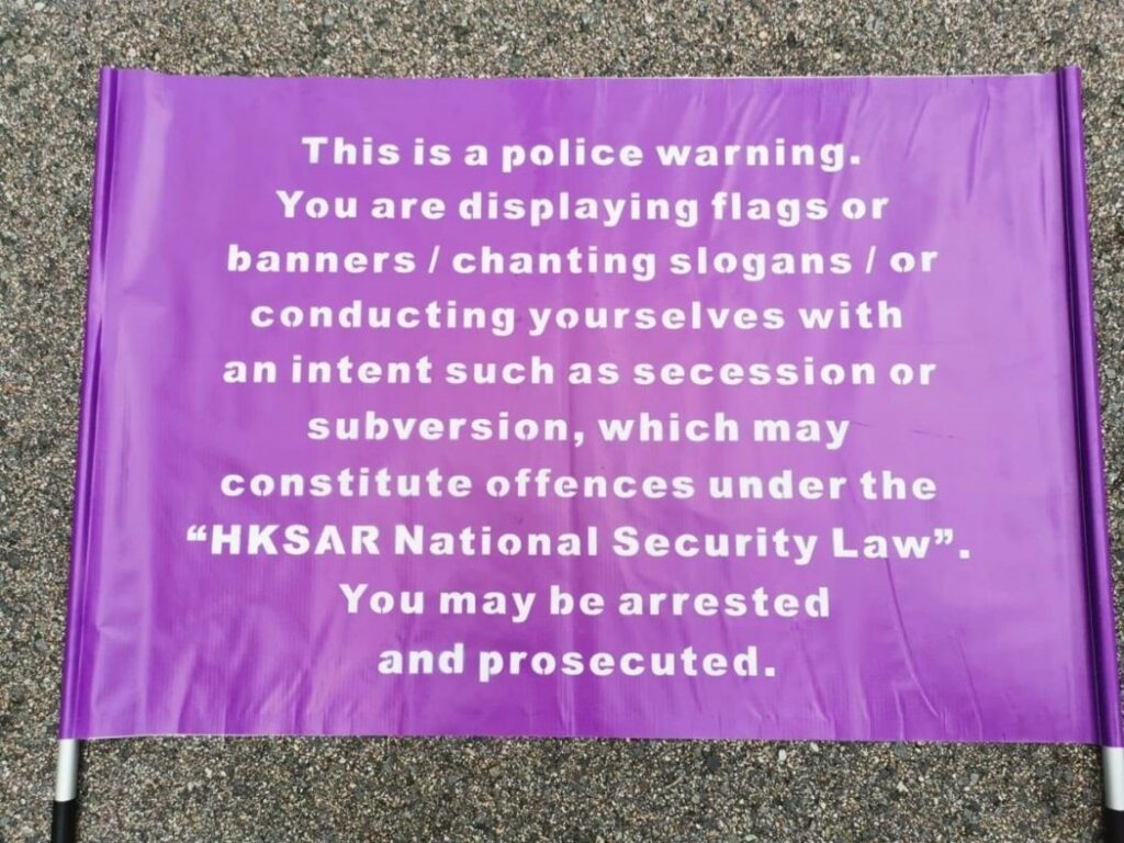 A purple banner that reads: "This is a police warning. You are displaying flags or banners/ chanting slogans/ or conducting yourselves with an intent such as secession or subversion, which may constitute offences under the "HKSAR National Security Law". You may be arrested and prosecuted.
