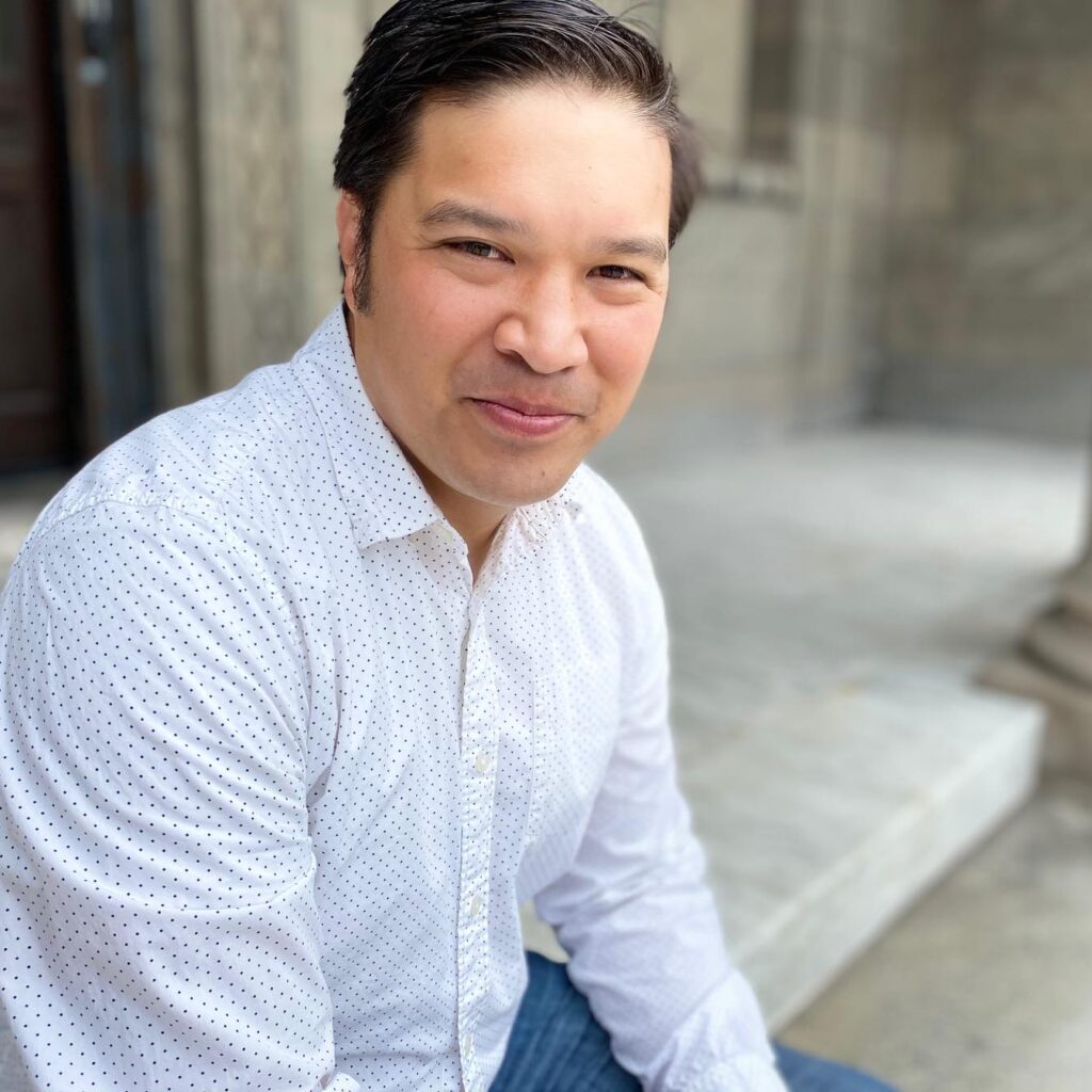 Photo of author sitting outside on concrete ledge, wearing a white button-up shirt with small polka dots