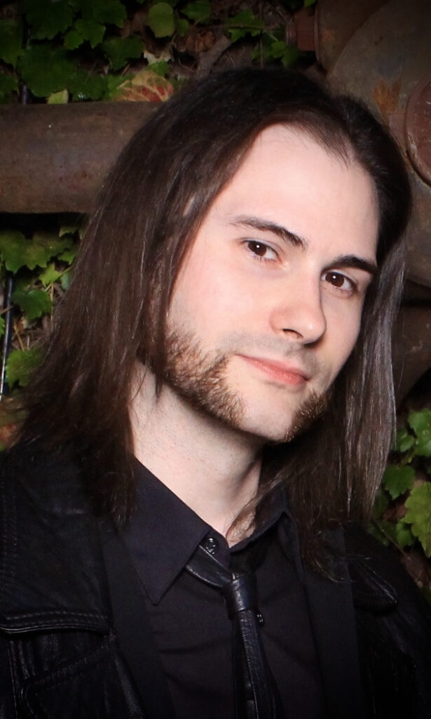 Photo of Sean Gill in a black shirt and jacket, posing in front of what appears to be a wall of plants. 