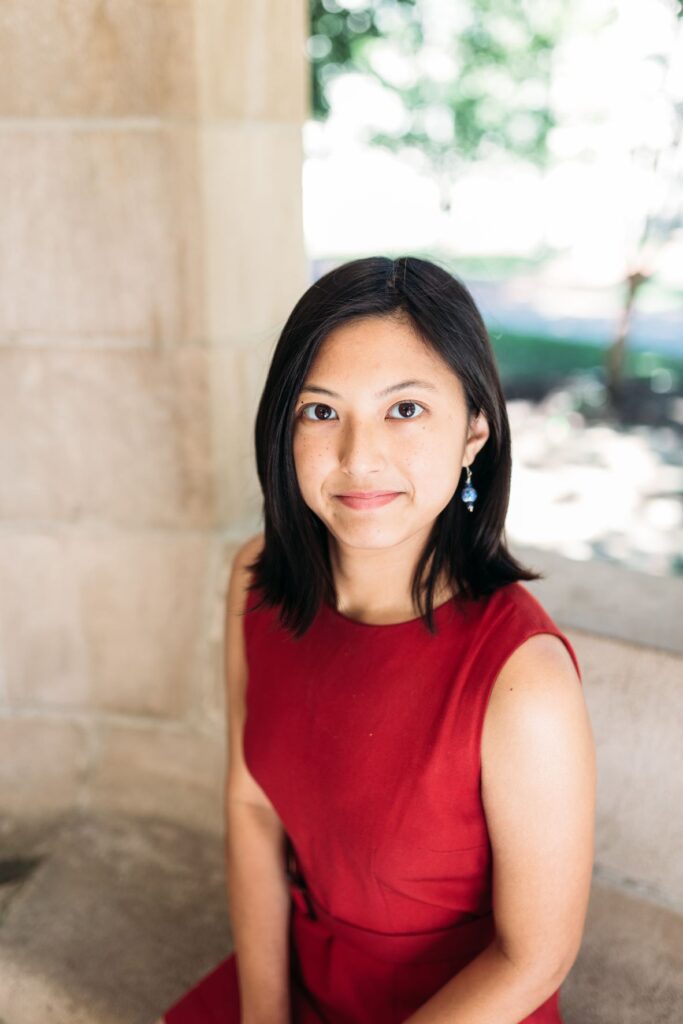 Photo of author wearing a red sleeveless dress, sitting in an outside structure whose windows open out to a blurry scene of trees. 