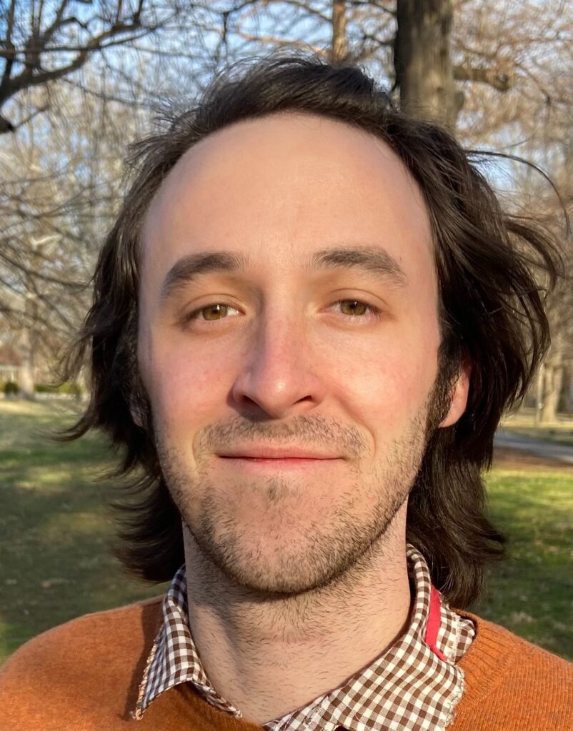 Elias Hutchinson in a park, wearing a brown-and-white checked shirt and orange sweater, smiling with his  mouth closed.