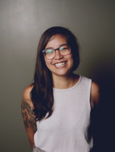 Photo of Danni Quintos with a gray background and the author smiling, wearing a white tanktop and glasses, and tattoos on her right arm.