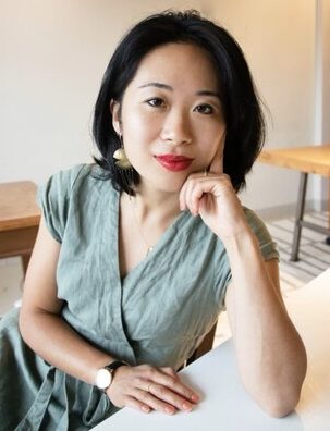 Author photo of Carlina Duan, leaning forward on a desk, wearing a green dress and gold-black watch.