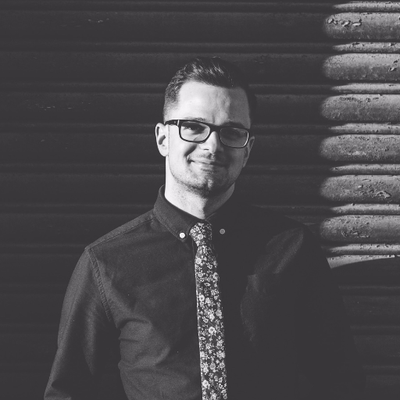 A black-and-white photo of Michael Alessi, in dark-rimmed glasses, wearing a shirt and tie, with log-style siding behind him.