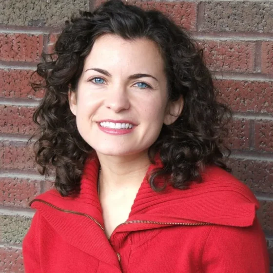 A woman with black, curly, shoulder-length hair and blue eyes, smiling. She stands in front of a brick wall, wearing a red coat.