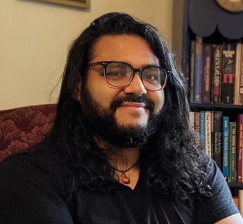 A man with long, wavy, black hair, brown eyes, and a beard. He sits in a brown armchair in front of a beige wall and a small bookshelf, wearing a black shirt, an orange pendant, and black, square-shaped glasses.