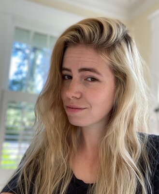 A woman with long, blond, wavy hair and dark eyes, smiling. She wears a black, long-sleeved shirt and stands in a white room with a window.