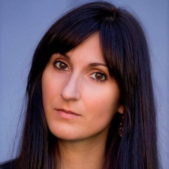 A headshot of a person with brown eyes and straight, dark hair and bangs that goes past their shoulders They stand in front of a blue background.
