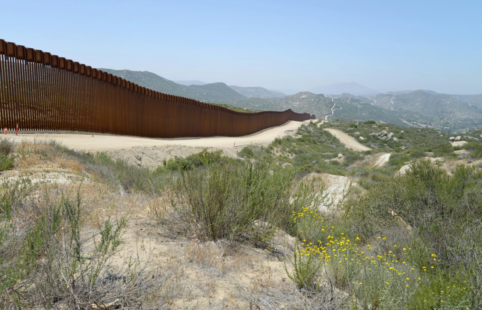 A photo of the Mexican-American border. A tall, metal fence stretches on for miles to the right. The terrain surrounding the fence is sandy, with dry plants and brittlebush.