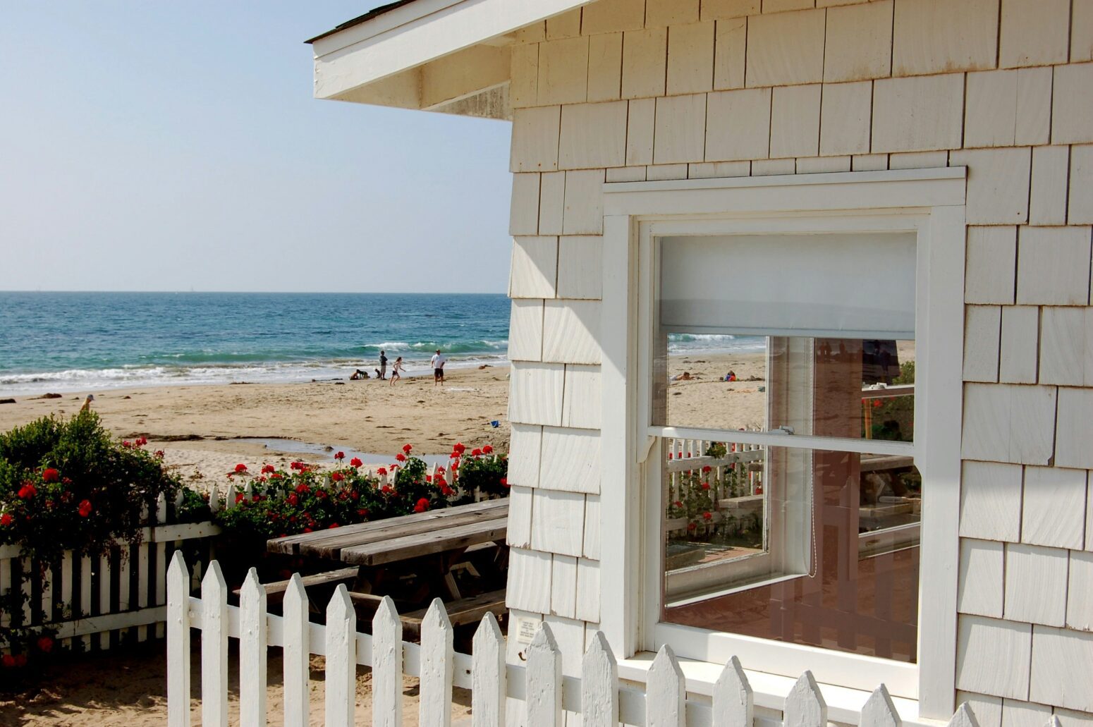 The corner of a white shingled house with a white picket fence and border of roses near it. In the distance people are on the beach near the ocean.