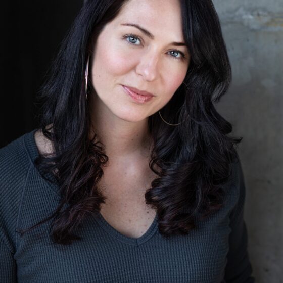 A woman with long, curly, black hair that goes down to her chest, tilting her head to the right and smiling. She stands in front of a gray wall, wearing a dark-gray, v-neck shirt.