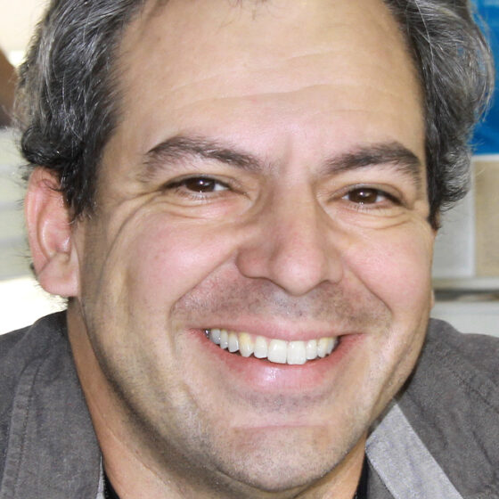 A man with gray hair, smiling. He wears a black top with a gray overshirt.
