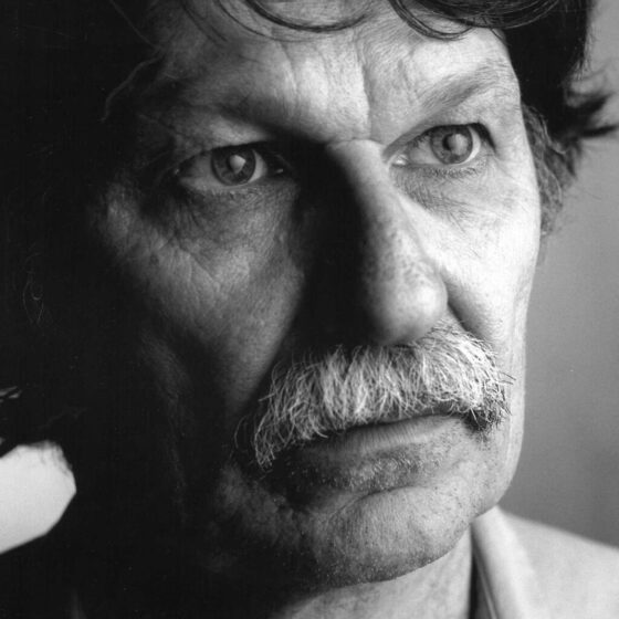 A grayscale photo of a man with dark hair and a lightly-colored mustache, looking to the right. He stands in front of a gray background, and wears a light-colored shirt.