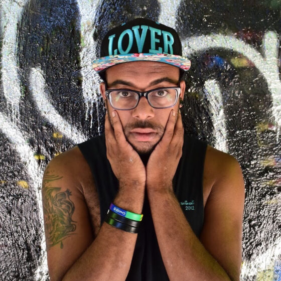 A person wearing a black hat that says "LOVER" in blue text, rectangular glasses, and a black tank top. They out their hands up to their cheeks and lean up against a wall covered in graffiti.
