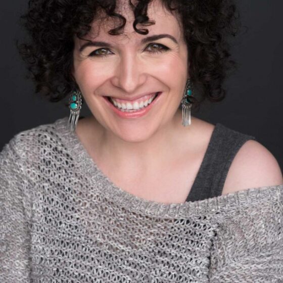 A white woman with dark curly hair. She's smiling and wearing an off-the-shoulder knit sweater over a tank top, and dangling earrings.