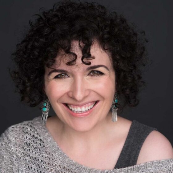 A white woman with dark curly hair. She's smiling and wearing an off-the-shoulder knit sweater over a tank top, and dangling earrings.