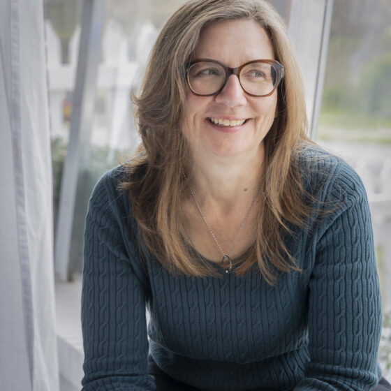 A woman with long, straight, gray-and-brown hair, smiling, looking toward the right, and putting her elbows on her knees. She sits in front of a large window, and wears a teal, long-sleeved shirt, black pants, black, cat-eye glasses, and a silver chain with a black ring on it.