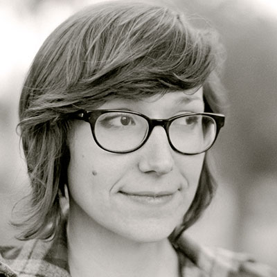 A grayscale photo of a woman with short, straight hair and dark eyes. She wears black, cat-eye glasses and a plaid shirt.