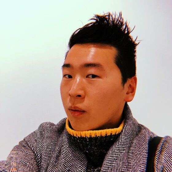 A person with short, spiky, black hair, facing towards the left. They are standing in front of a white background, wearing a black-and-yellow, turtleneck sweater underneath a black and white cardigan.