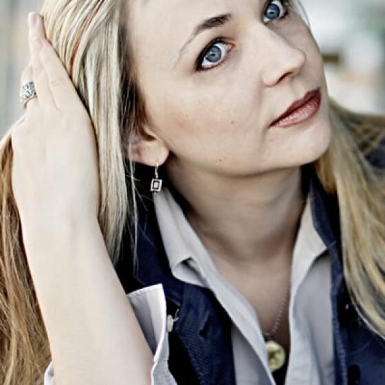 A woman with long, blonde hair to her chest, running her right hand through her hair and tilting her head up and to the right. She wears a white, button-down shirt and a blue blazer.