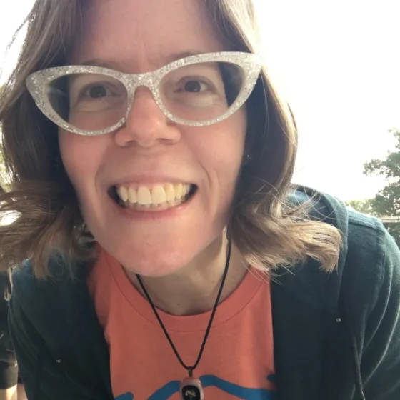 A woman with wavy, brown hair and brown eyes, leaning over to the camera, smiling. She stands outside wearing an orange shirt, a black hoodie overtop, a black necklace, and white, cat-eye glasses.