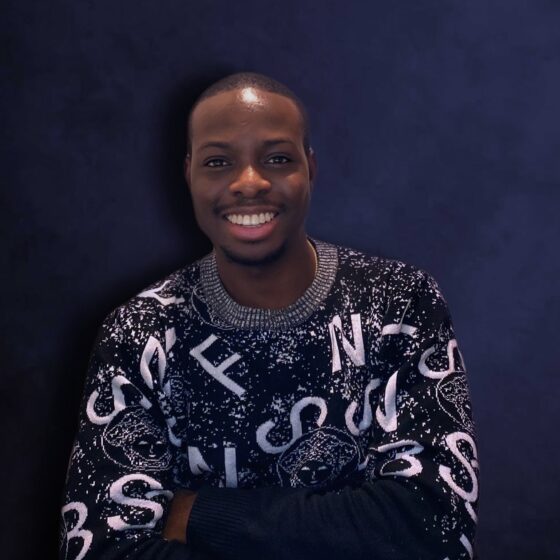 An African man smiling and wearing a sweater with gray and white letters on it. He's in front of a navy blue background