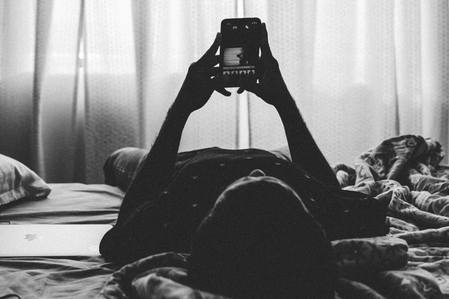 A grayscale photo of a person with dark hair, laying in bed and on their phone. They lay next to a laptop, and in front of a large window covered by curtains.