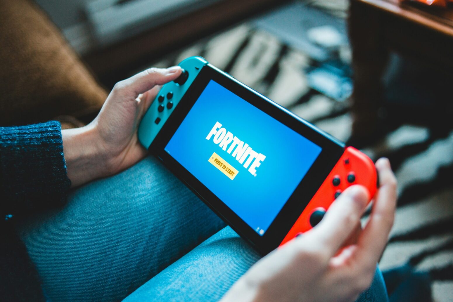 The view of a person's hands holding a Nintendo Switch on the Fortnite start-up screen. The person wears jeans. The room they are in has a zebra-print rug on the ground.