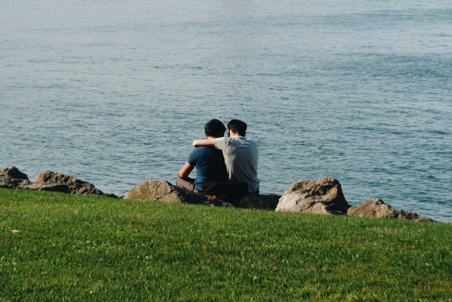 Two men sitting on the rocks next to a lake. They are embracing, and looking out at the water.