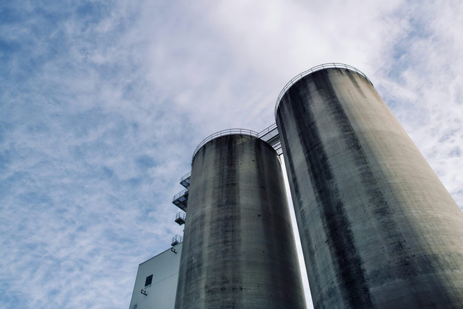 A view looking up at two tall grain silos against a blue, cloudy sky. They are light gray with runoff stains, and a small bridge connects between them on the top.