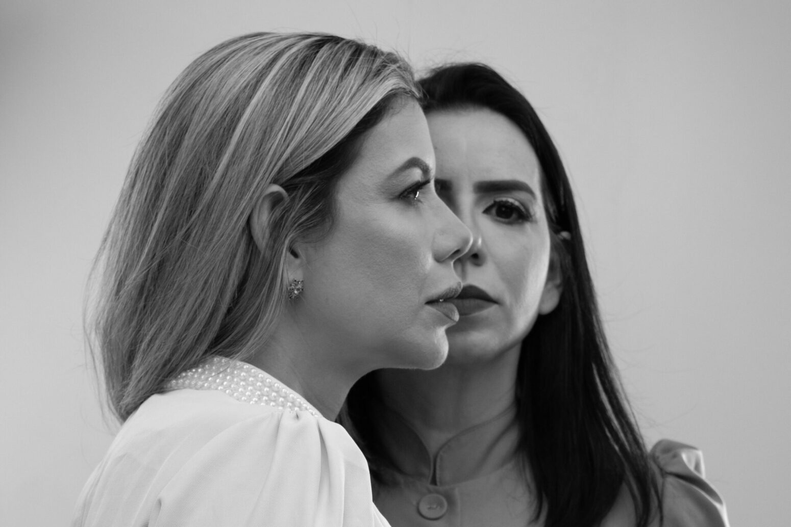 A grayscale photo of two women from the shoulders-up. One is facing forward, with long, dark, straight hair and wearing a blouse with a button. The other stands in front of the first woman, partially covering her. She has long, light, straight hair, wears a white blouse with pearls around the collar, and is completely facing the right. Both women have blank faces, and the background is a plain gray.