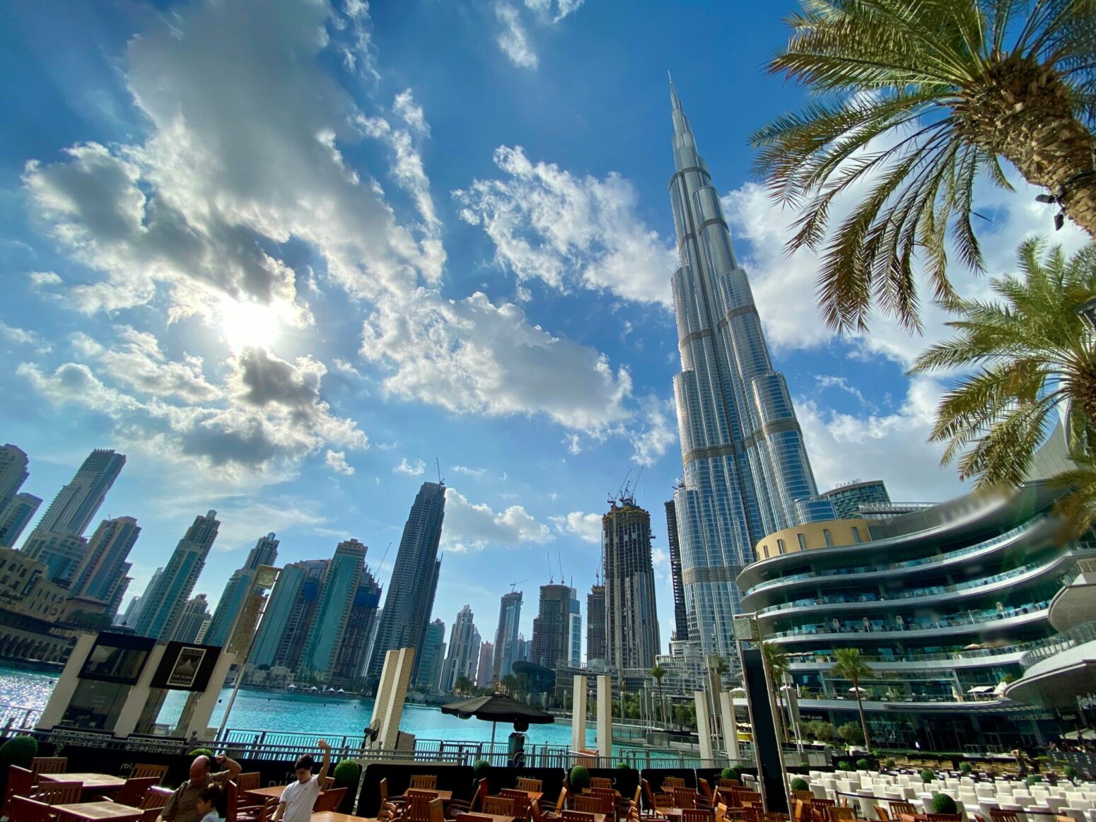 The cityscape of Dubai, with Burj Khalifa on the right. There are tall skyscrapers against a bright-blue, cloudy sky. Water reflects the blue of the sky. Palm trees grow on the right.