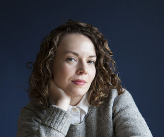 Bess Winter, a white woman with curly brown hair, wears a gray sweater and white collared shirt and sits at a wooden table in front of a blue background.