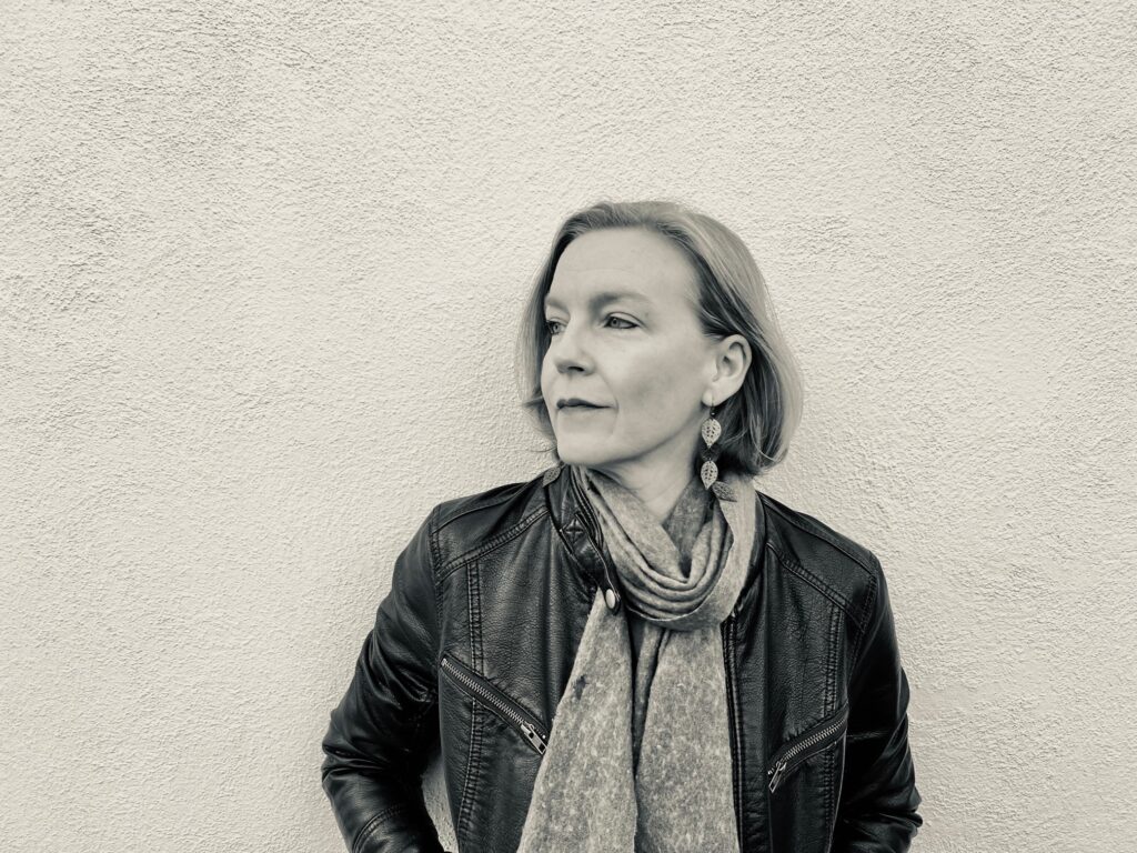 Leslie Parry wears a short bob, a black leather jacket, and a light scarf. She looks off into the distance as she stands in front of a white background.