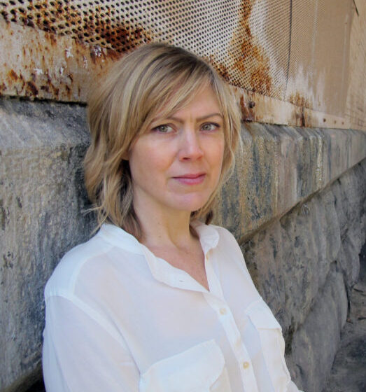 A woman with short, straight, blonde hair and bangs, leaning up against a stone-and-brick wall. She is wearing a white, button-down shirt.