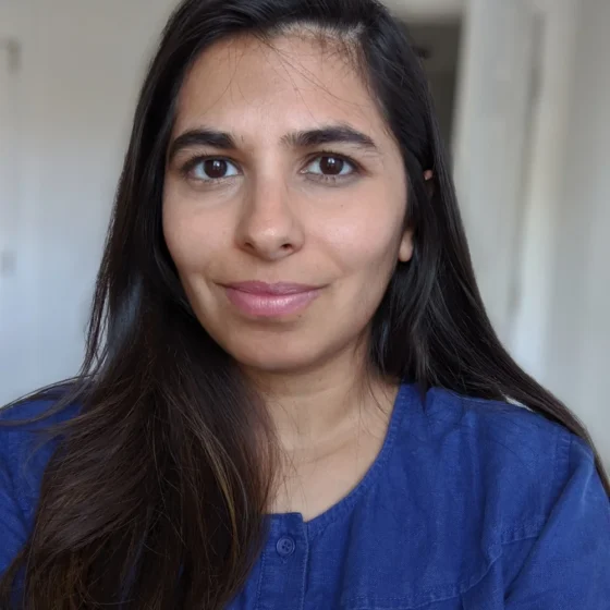 A woman with long, straight, black hair, looking forward and smiling. She is standing in a room with white walls, and wearing a dark-blue shirt.