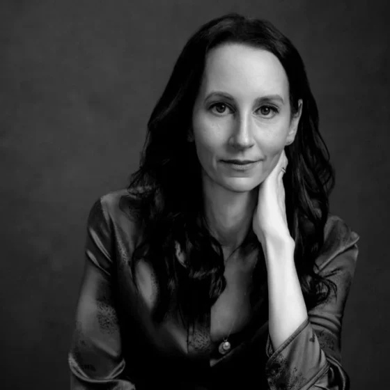 Black and white photo of a white woman with long hair. She's wearing a shiny dark-colored blouse, and leaning her chin on her left hand.