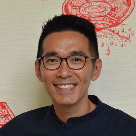 O Thiam Chin, an Asian man with a cropped haircut and horn-rimmed glasses, smiles in front of a red-and-white toile-like background.