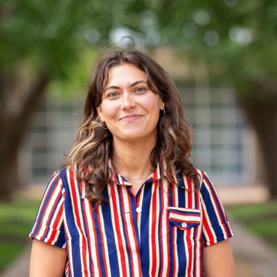 A woman with long, curly, brown hair with blonde highlights, smiling. She is standing outside on a path with trees on either side, and she is wearing a button-up t-shirt with blue, white, yellow, and red stripes on it.