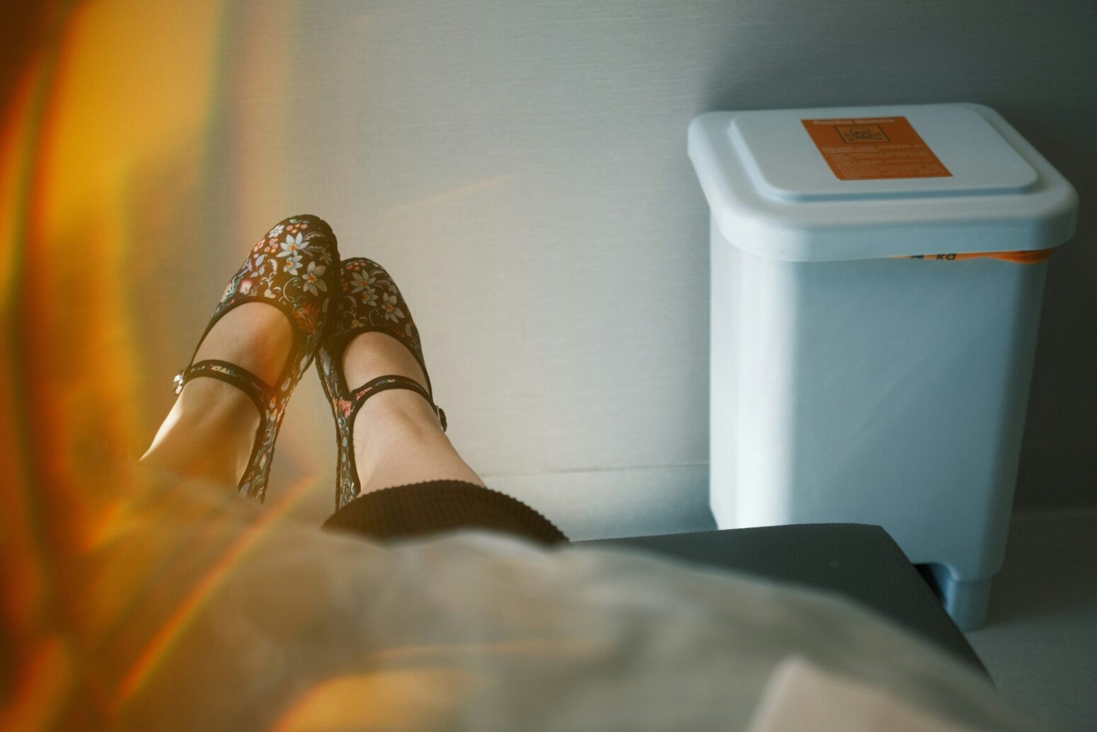 A perspective shot of a woman looking at her shoes. They are black flats with little flowers embroidered on them. She sits in a hospital room, with a trash can marked as a biohazard against the white wall opposite her.
