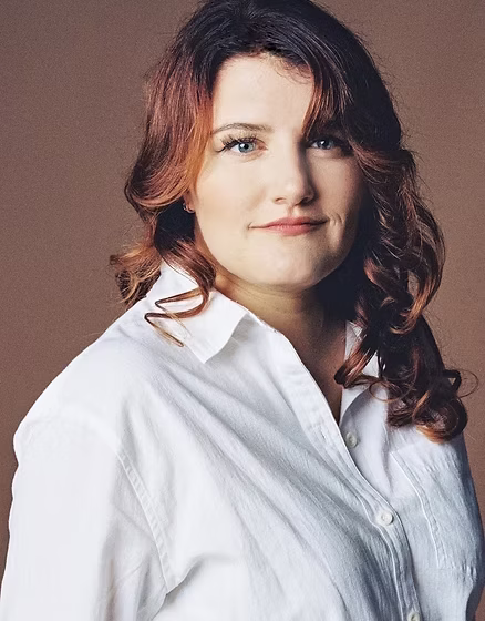 A white woman with brown/red hair past her shoulders, curled. She's wearing a white button-down collared shirt and standing in front of a brown background