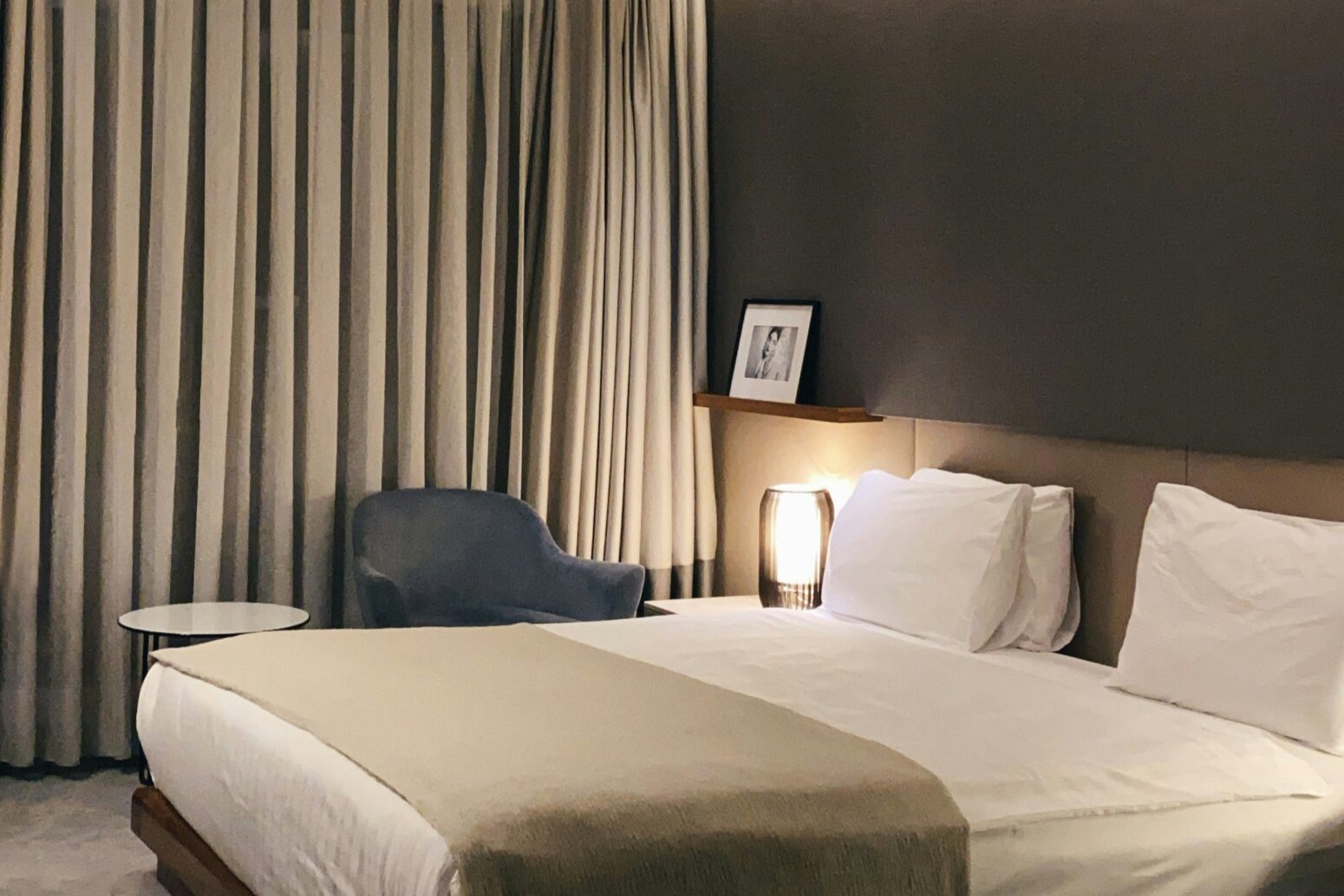 A hotel room with dark-gray walls and a light-gray carpet. A bed with white sheets and a gray blanket sits in the middle. A gray chair with a white side table sits in from of curtains that cover one wall.