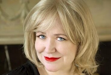 Kimberly Elkins wearing a black textured thin sweater and black shirt, and red lipstick, and smiling.