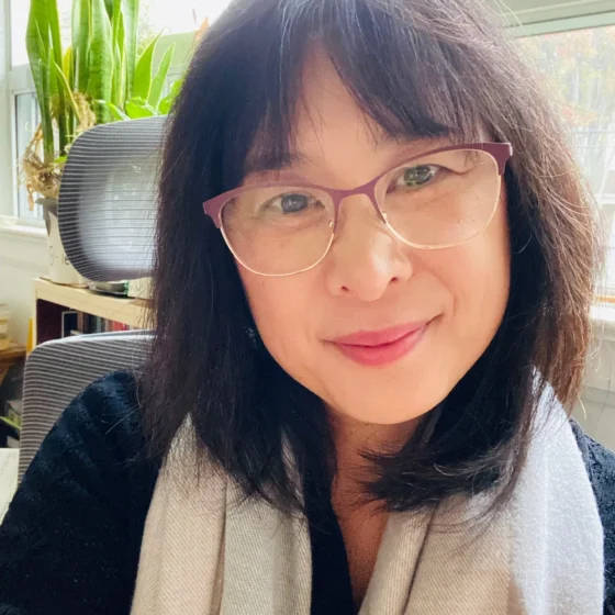 A woman with straight, black hair with bangs, smiling. She is sitting in a brightly-lit office, and wearing a black shirt, a gray scarf, and round glasses.