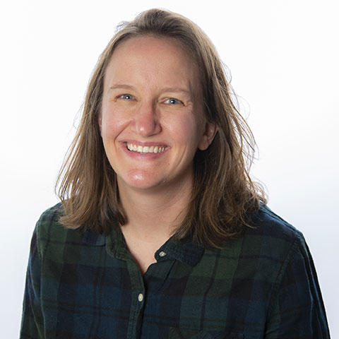A woman with straight, brown hair, just past her shoulders, smiling. She stands in front of a white background, and wears a dark, green-and-black, plaid shirt.